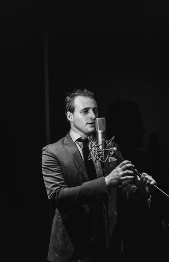 Black-and-white shot of a singer in a suit and tie recording vocals at a studio microphone