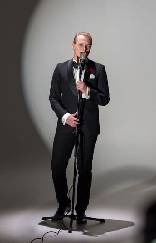 Singer in a tuxedo with bow tie performs at a vintage microphone under a round spotlight