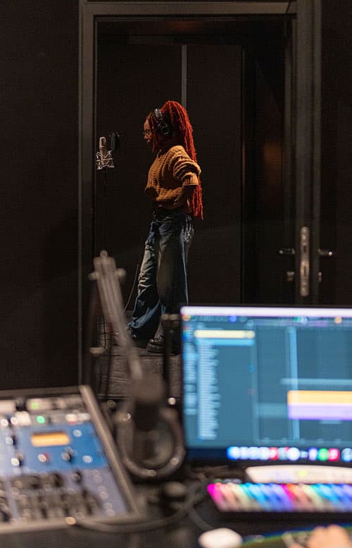 Vocalist with long red locs and headphones records in the booth, seen past the control room gear