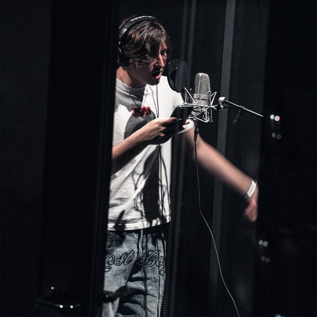 Young singer in headphones reads lyrics from a phone while recording at a studio microphone