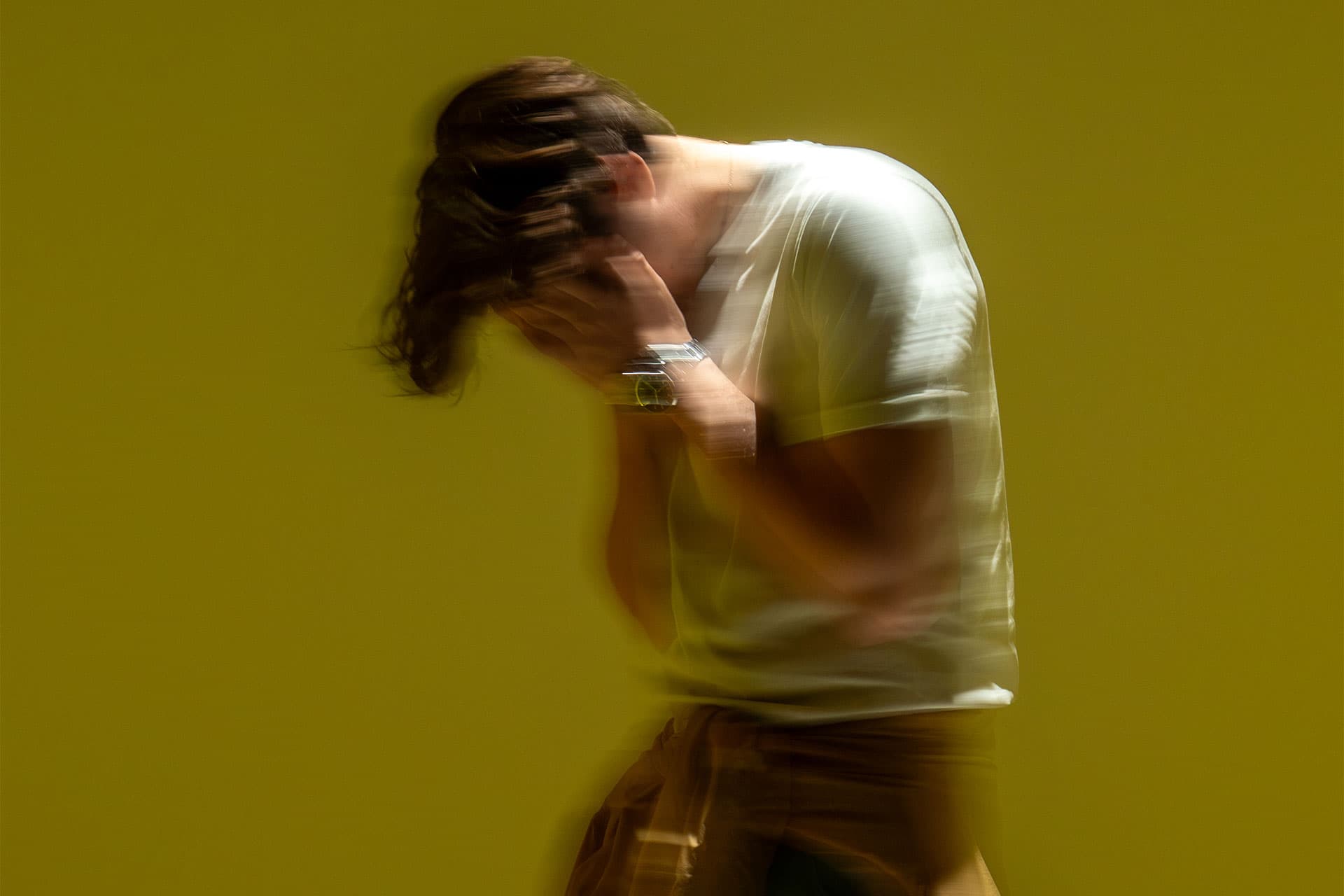 Motion-blurred figure in a white T-shirt clutches his head against a mustard-yellow background
