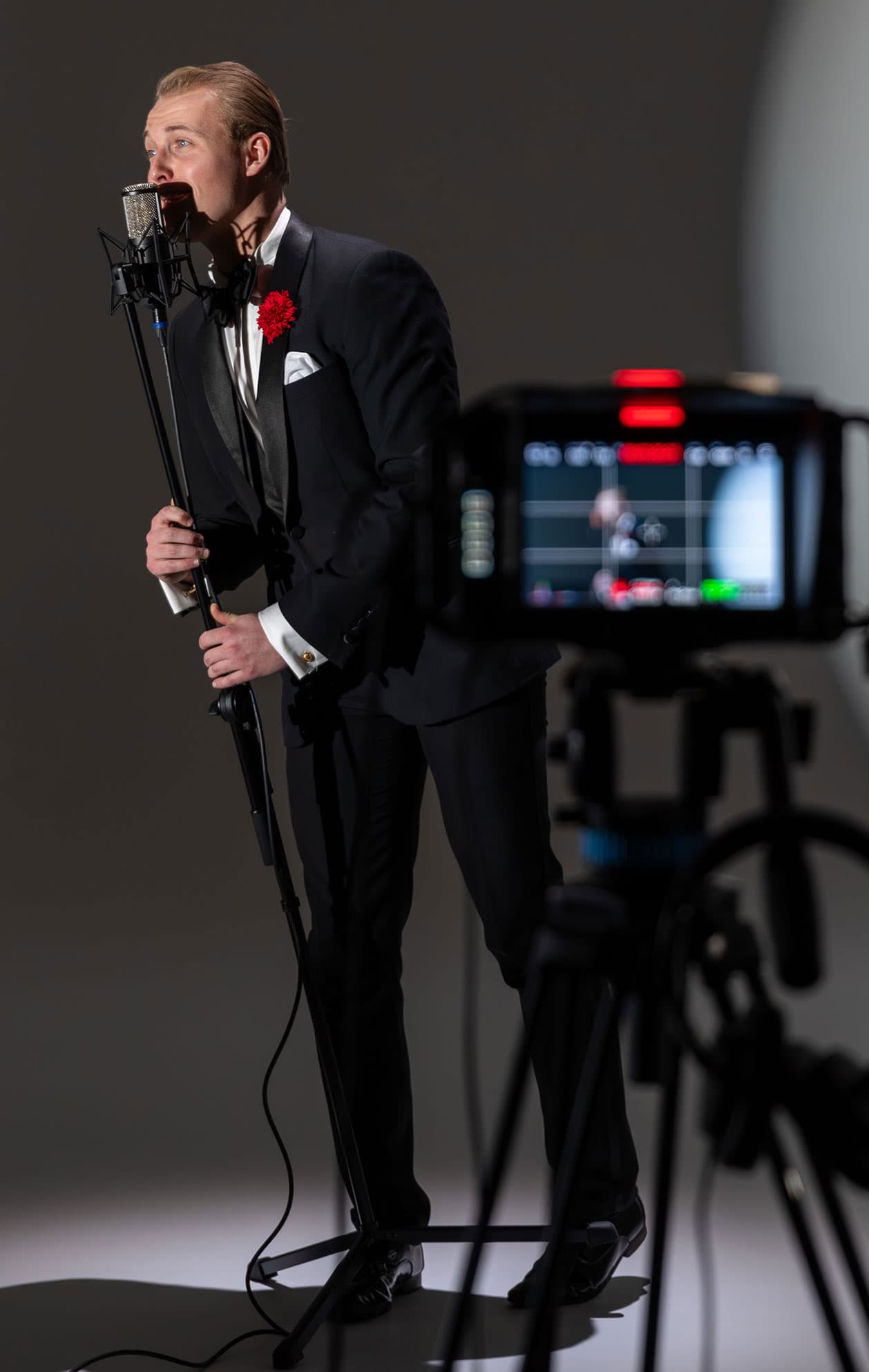 Singer in a tuxedo with a red boutonniere performs at a microphone as a video camera films him
