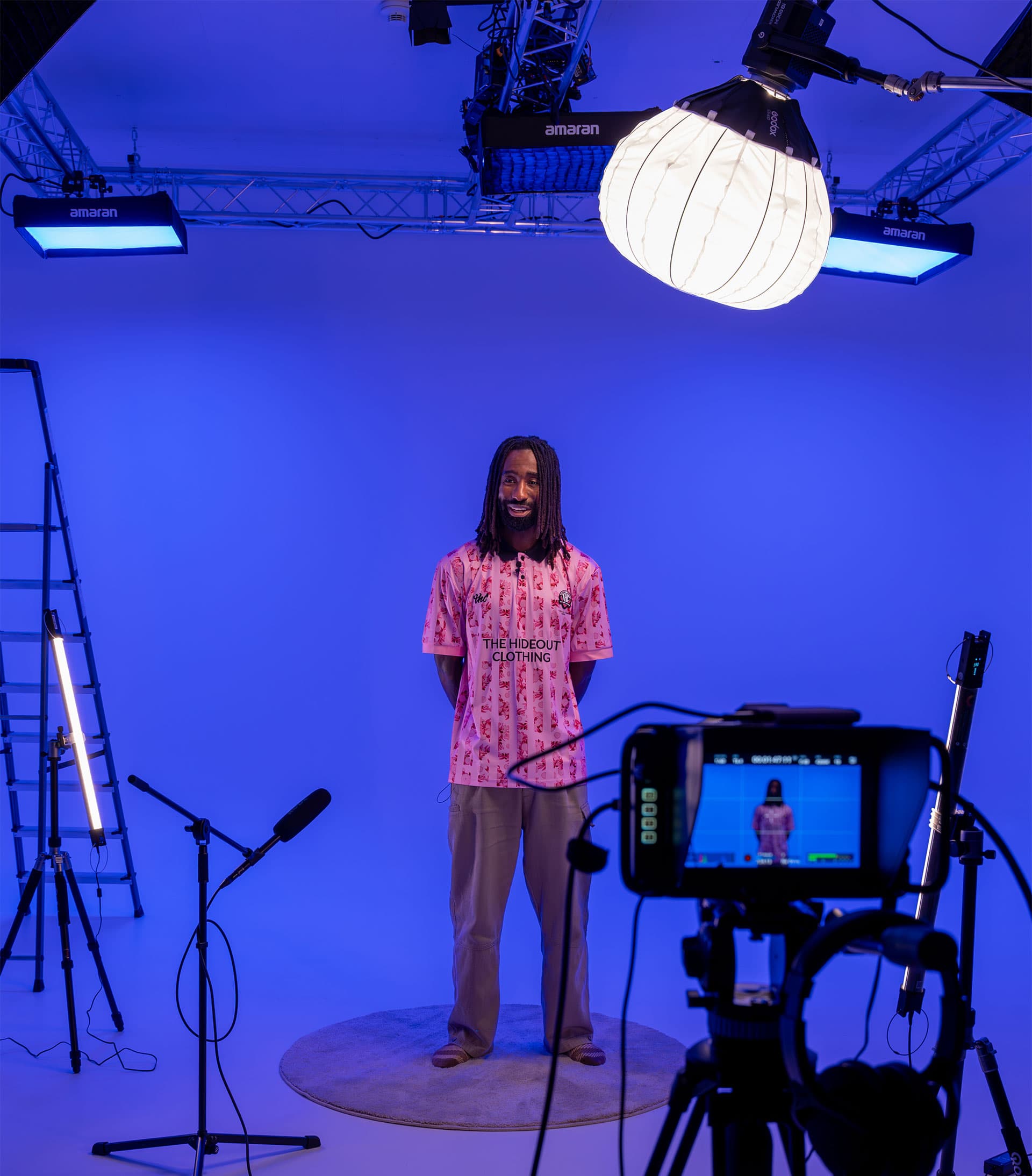 Man with dreadlocks in a pink patterned shirt stands on a blue-lit cyclorama among studio lights