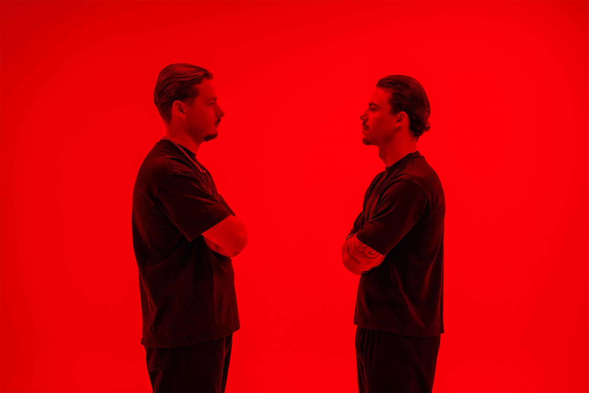 Two men in black stand face to face in profile against an intense red backdrop