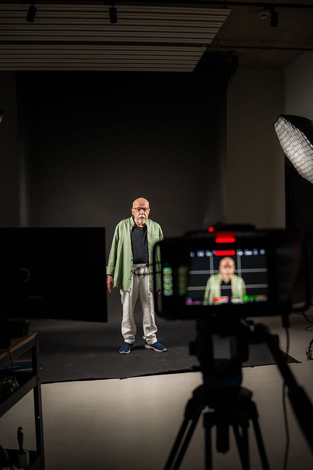 Paolo Coelho standing on a dark studio backdrop, framed on a camera monitor in the foreground