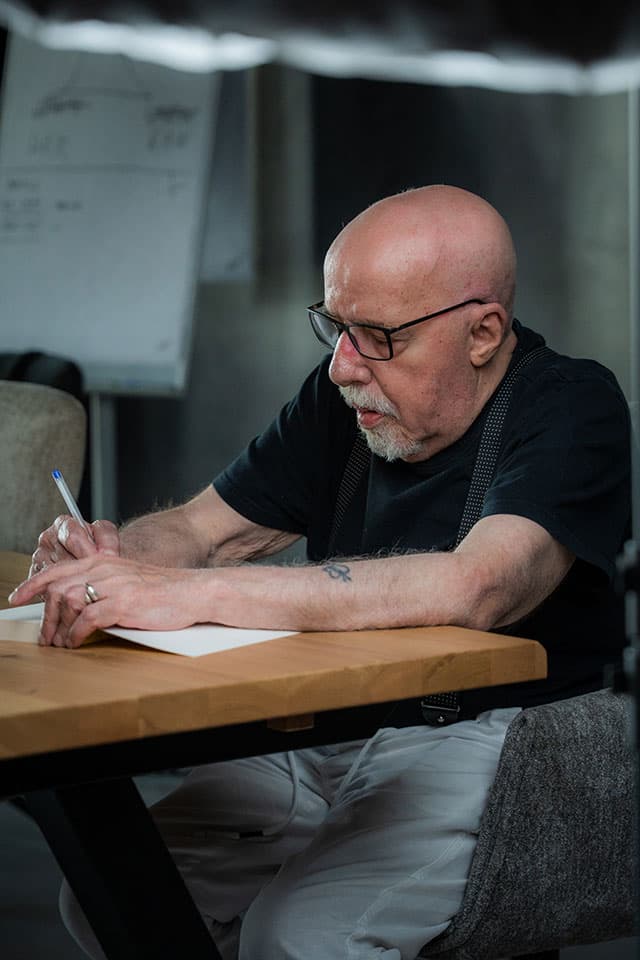 Paolo Coelho seated at a wooden table, writing on a sheet of paper with a pen