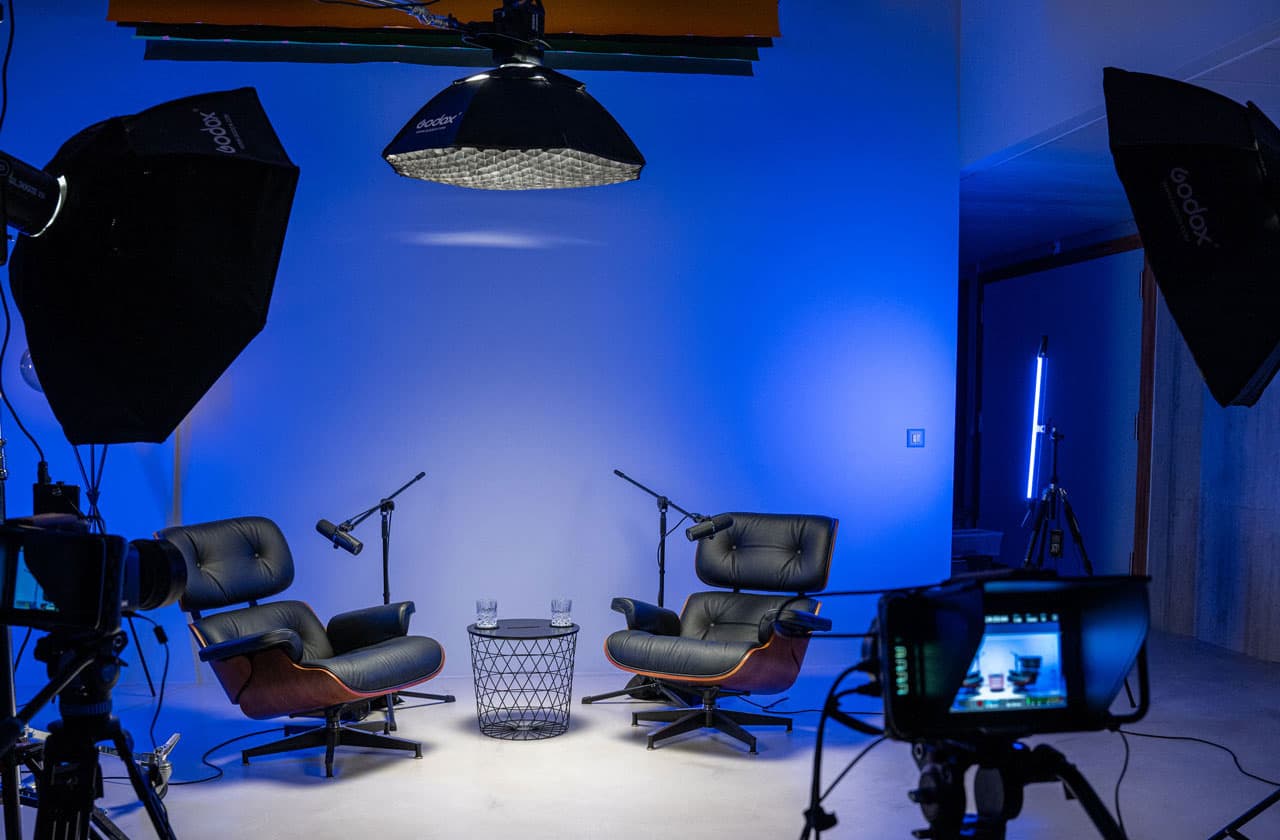 Plateau de podcast vide avec deux fauteuils en cuir, micros, softbox et caméra dans un studio éclairé en bleu