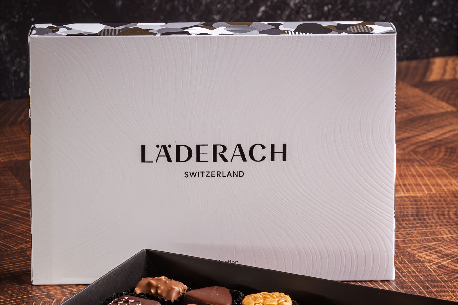 White gift box of chocolates standing on a wooden table, assorted pralines in an open tray in front