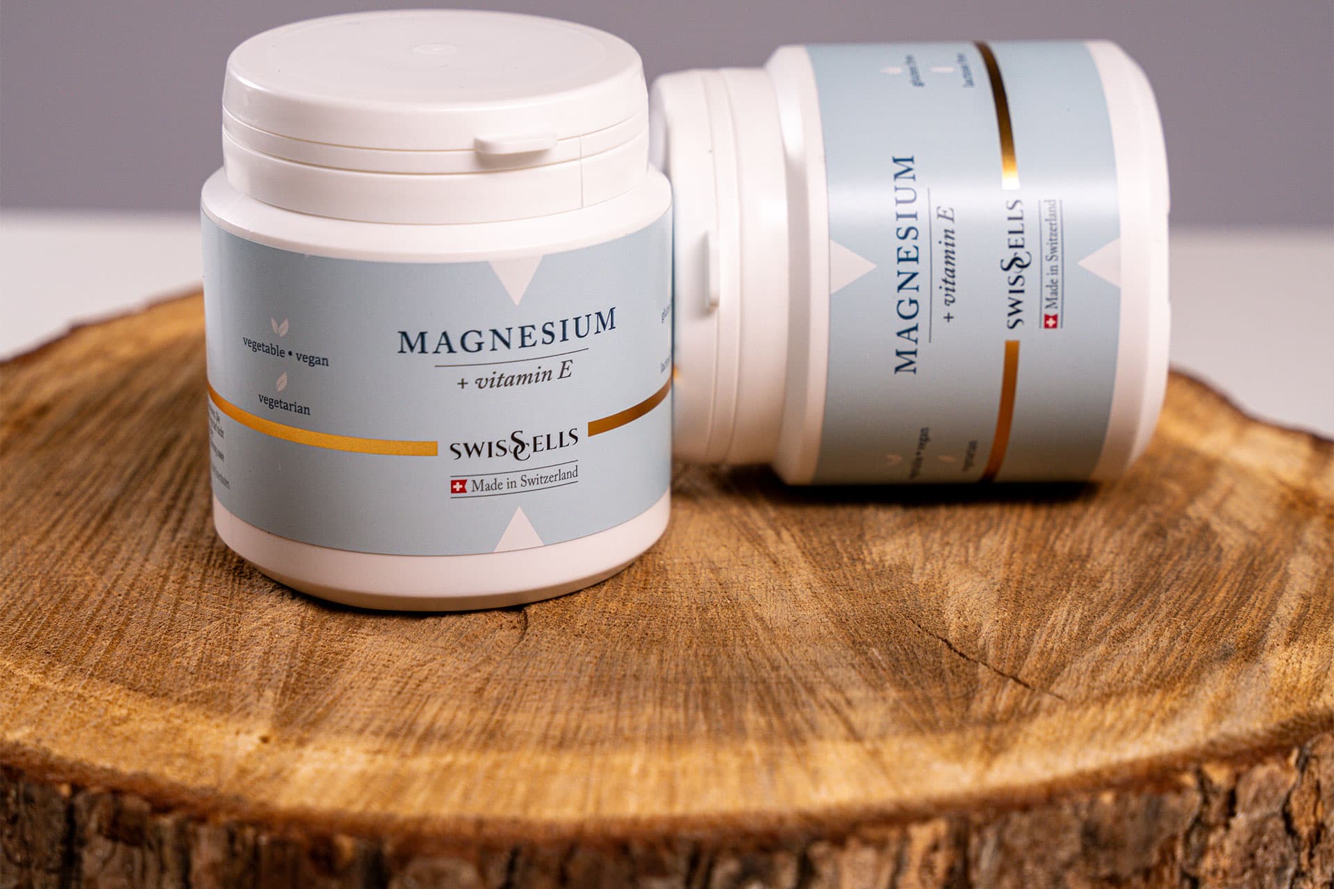 Two white jars of magnesium supplements with pale blue labels on a wooden log slice, one lying on its side
