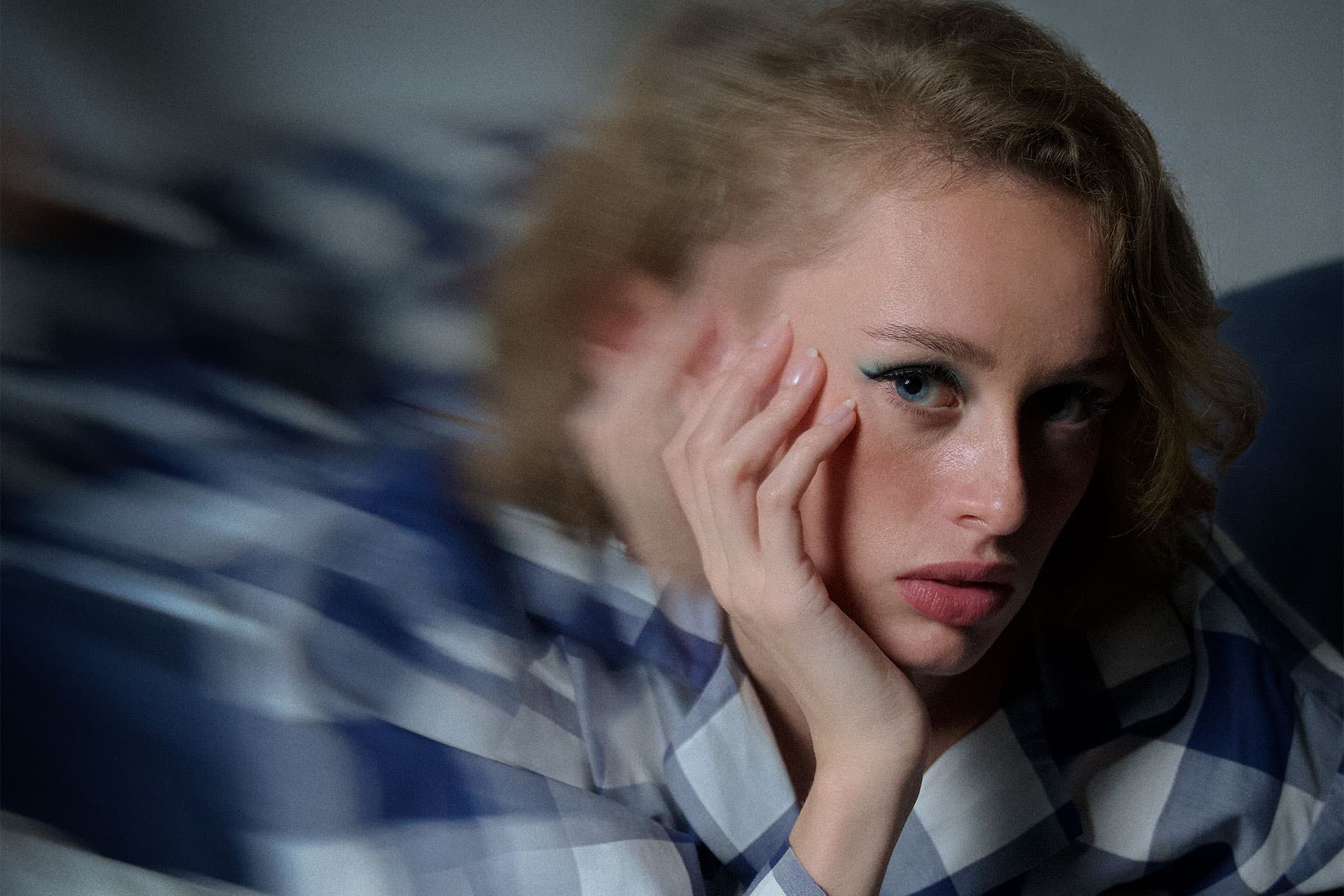 Femme au fard à paupières bleu, la joue dans la main, en pyjama à carreaux bleu et blanc, literie floue autour