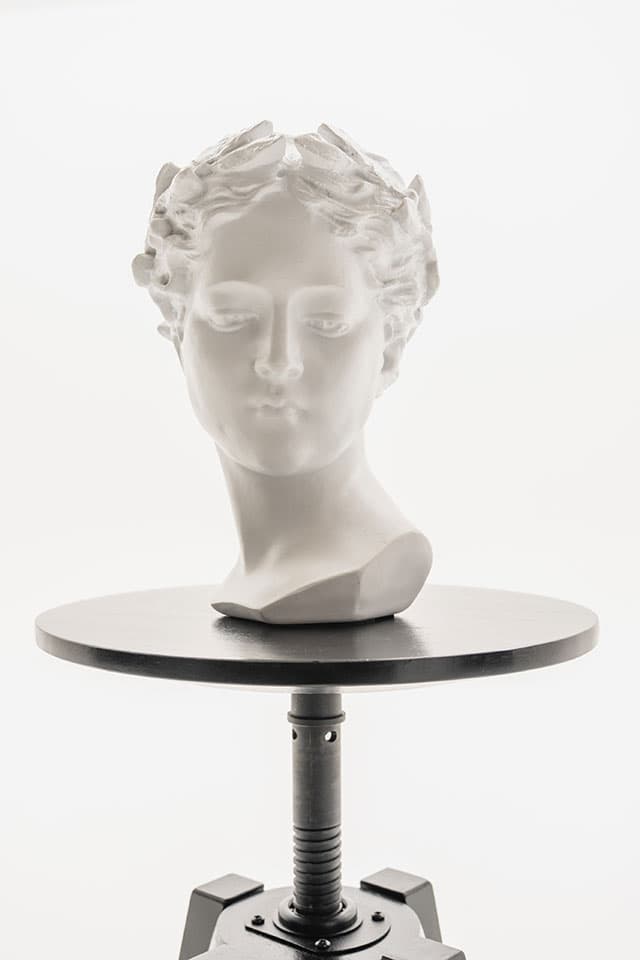 White plaster head with laurel wreath on a round black stool against a bright white background
