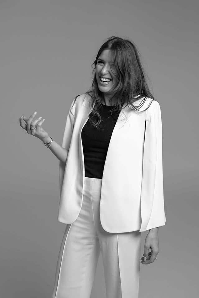 Black-and-white portrait of a laughing woman in a white cape blazer and trousers on a grey studio backdrop
