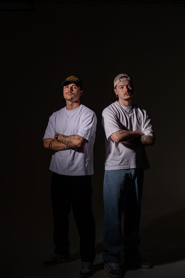 Two men in caps and white t-shirts standing with arms crossed under dramatic low light on a dark backdrop